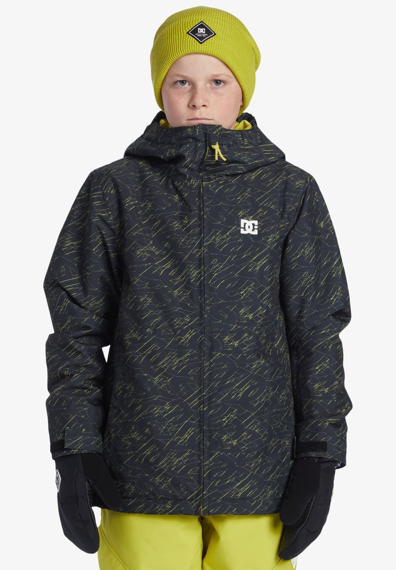 Svart och gula mönstrade snöjacka med huva, framdragkedja och synlig DC-logga. Bärs med en matchande gul stickad mössa och svarta handskar.