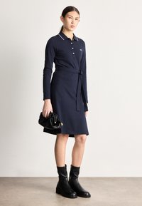 Tumši zila polo kleita ar garām piedurknēm un jostu viduklī, ar apkakli baltā apmalē un nelielu logotipu. Kombinēta ar melniem potīšu zābakiem un rokassomu.