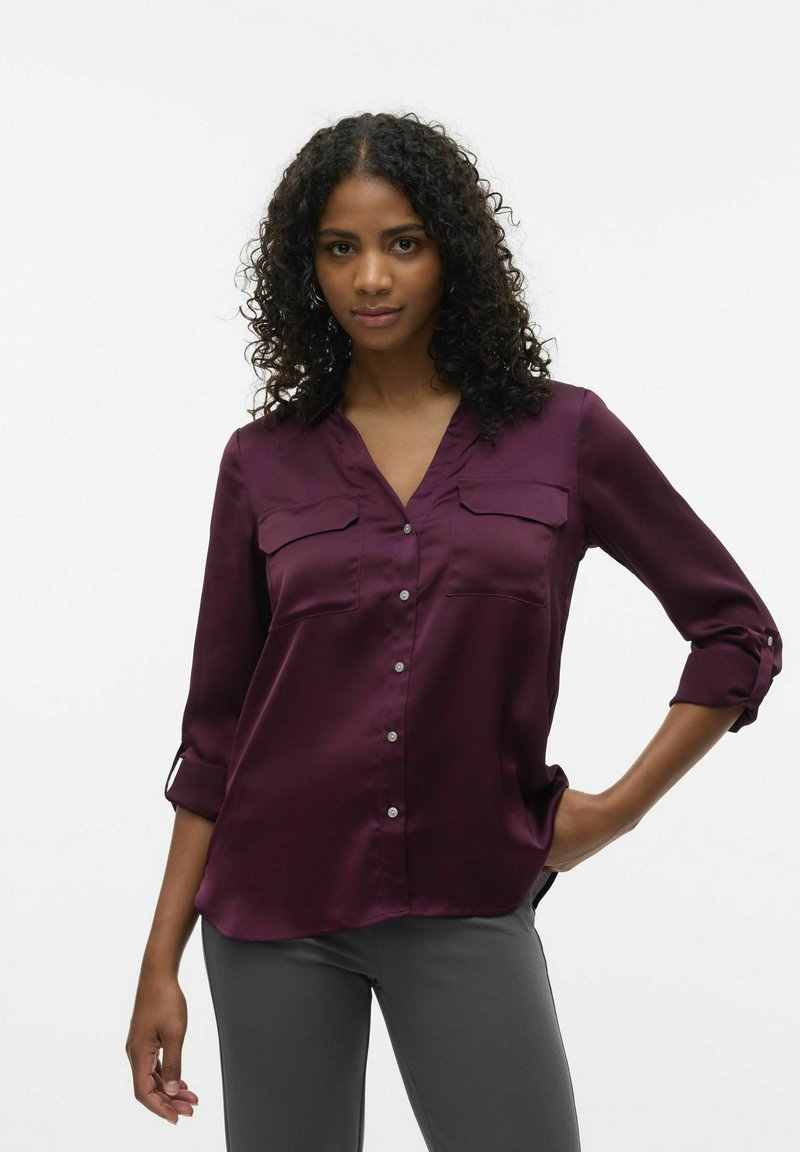 Chemise en soie bordeaux avec un décolleté en V, deux poches poitrine et des manches trois-quarts. Présente une fermeture à boutons et une texture lisse et brillante.