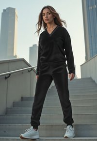 Zwart, zacht stof loungewear set met een V-hals trui en bijpassende taps toelopende broek. Voltooid met witte sportschoenen en een gladde afwerking.