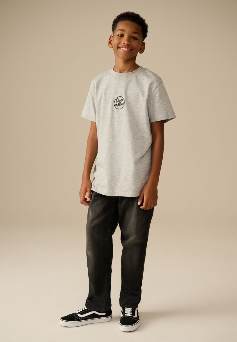 Dreng står og smiler iført en grå t-shirt med tekst, sorte jeans og sorte og hvide sneakers på beige baggrund.