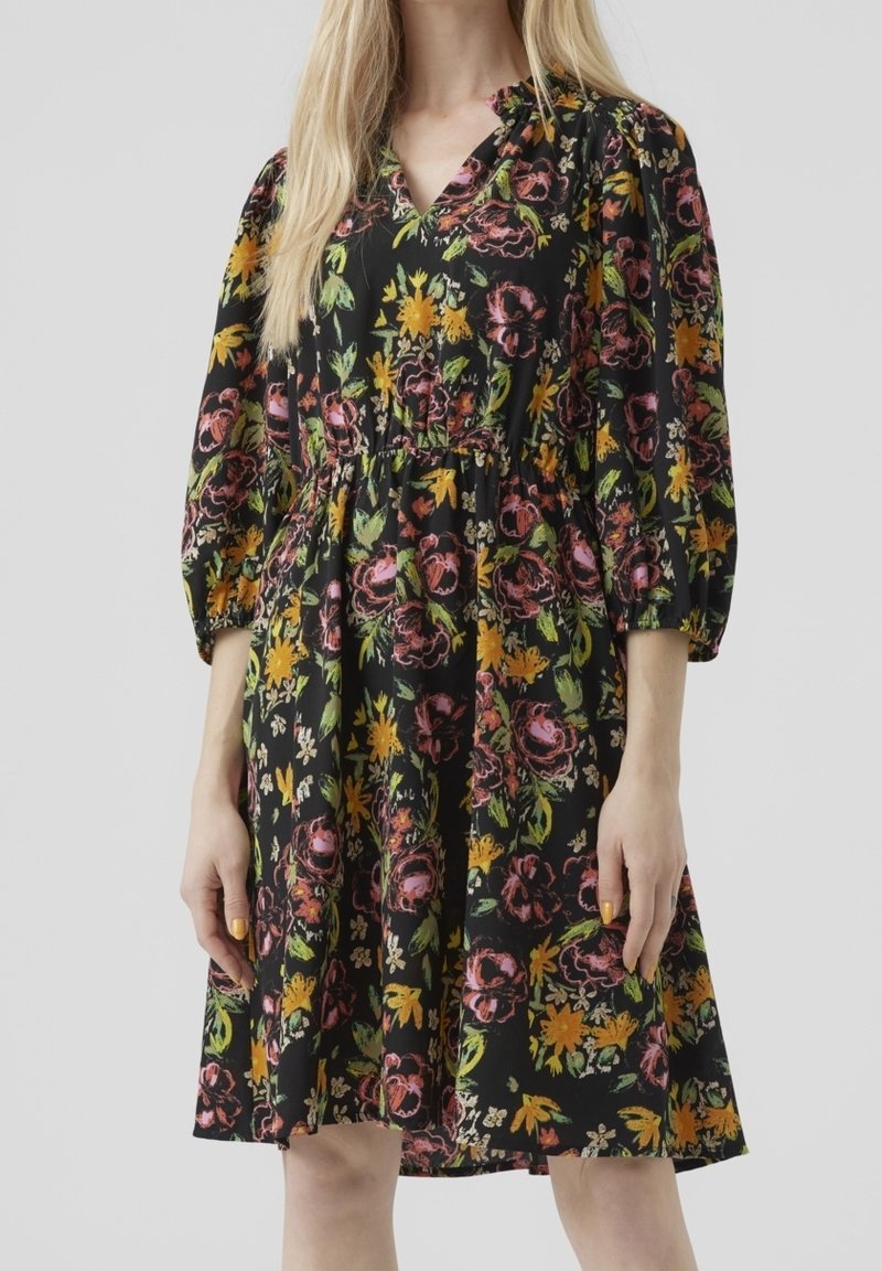 Femme portant une robe noire longueur genou avec un imprimé floral rose, jaune et vert, manches bouffantes trois-quarts, sur fond gris.