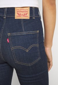 Jeans de mezclilla azul oscuro con un diseño clásico de cinco bolsillos. Presentan un parche de cuero con texto rojo en la cintura y acentos de costura amarilla.