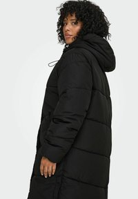 Melnā pūkaina jaka ar kapuci, izceļot quiltotas daļas, pilna garuma dizainu un sānu kabatas. Materiāls šķiet gluds un izturīgs.