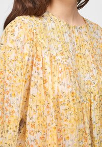 Blusa floreale leggera con tessuto a motivo giallo e pastello. Presenta plissettature, un collo rotondo e delicati accenti floreali.