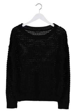 Person, der går, iført en sort strikket sweater, mørke skinny jeans, sorte ankelstøvler og en brun crossbody taske på en lys baggrund.