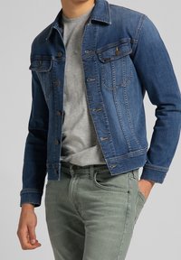 Jeansjacka i mörkblått med en klassisk design, försedd med två bröstfickor, silverfärgade tryckknappar och en strukturerad yta. Bärs över en grå t-shirt.