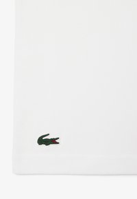 Camisa polo de algodón blanca con un logo de cocodrilo verde en el dobladillo inferior. La tela tiene una textura suave y un diseño de cuello clásico.