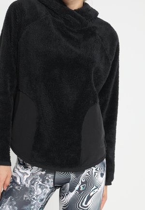 Frau, die einen schwarzen flauschigen Langarm-Pullover und abstrakte schwarz-weiße gemusterte Leggings trägt und vor einem schlichten Hintergrund steht.