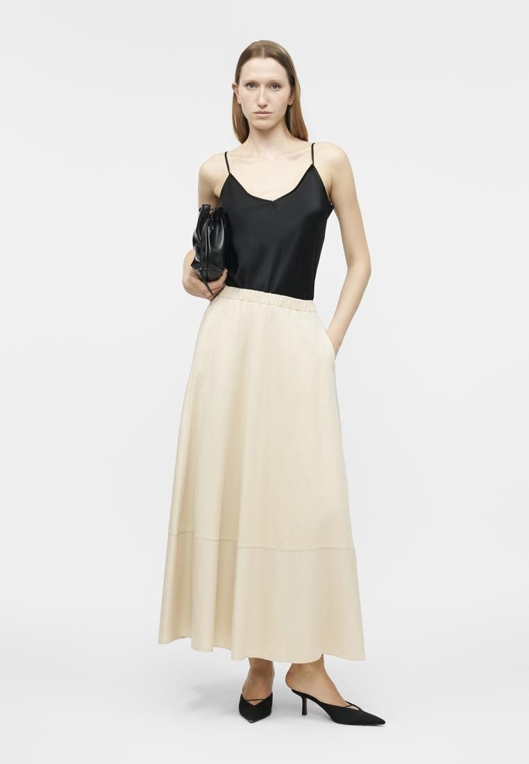 Femme portant un haut sans manches noir, une longue jupe beige, des talons noirs, tenant un sac à main noir, debout devant un fond clair uni.