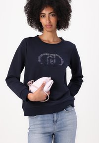 Felpa blu navy con colletto a girocollo e un accento di logo scintillante. Abbinata a jeans blu chiaro e a una piccola pochette rosa. Texture liscia su tutta la superficie.