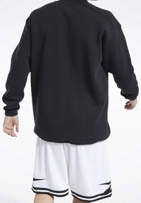 Persona vista de espaldas, vestida con una camiseta negra de manga larga y pantalones cortos de malla blancos con detalles negros, de pie contra un fondo blanco.