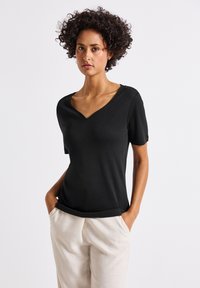 Černý krátký rukávový top s výstřihem do V a hladkým povrchem, v kombinaci se světle béžovými kalhotami. Modelka pózuje s rukama v kapsách.