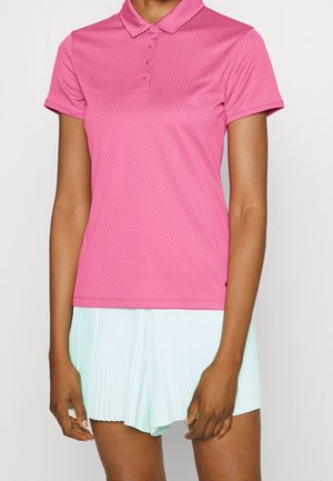 Vrouw draagt een roze textuur polo met korte mouwen en een lichtblauwe geplooide rok, staand tegen een effen lichte achtergrond.