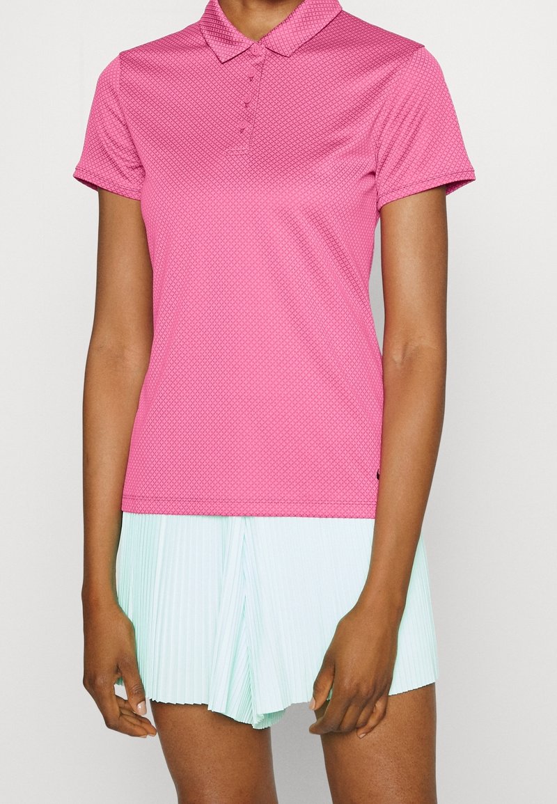 Femme portant un polo rose texturé à manches courtes et une jupe plissée bleu clair, debout devant un fond clair uni.