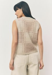 Kvinde med langt mørkt hår iført ærmeløs beige ternet strikvest og lysebeige bukser, vendt for at vise ryggen og sideprofilen.