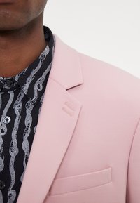 Blazer rose clair avec une texture lisse, doté d'un col cranté et de détails de couture subtils sur le revers. Associé à une chemise noire à motifs.