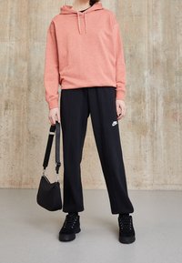 Korallfärgad hoodie med dragsnören, tillsammans med svarta Nike sweatpants. Svart handväska, platåskor med texturerad yta, stående på ett slätt golv.