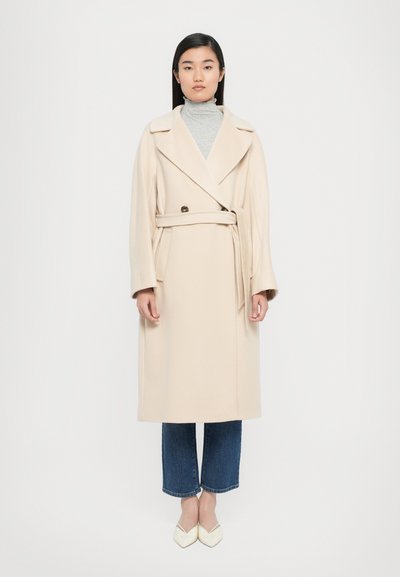 Cappotto lungo in misto lana color crema con ampi revers, una cintura in vita e due bottoni. Indossato sopra un dolcevita grigio e jeans blu.