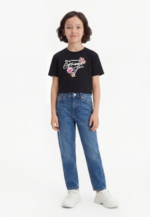 Enfant debout portant un t-shirt court noir à logo floral, jean taille haute bleu et baskets blanches épaisses sur fond blanc.