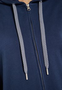 Sudadera con cremallera color azul marino que presenta un tejido texturizado, cremallera metálica y cordones trenzados blancos con puntas metálicas.