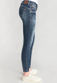Vaqueros ajustados de denim azul oscuro con secciones desgastadas, costuras visibles y detalles desgastados. Combinados con zapatillas blancas.