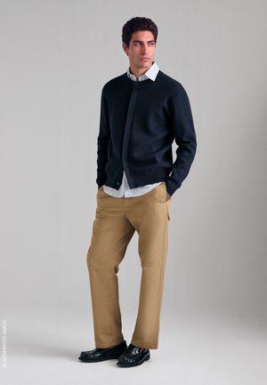 Uomo in piedi con le mani nelle tasche che indossa un cardigan nero, una camicia bianca abbottonata, pantaloni beige e scarpe nere su uno sfondo semplice.