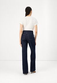 Frau mit geflochtenem Haar, die ein weißes T-Shirt, dunkle, hoch taillierte, gerade Jeans und braune Absätze trägt, steht vor einem schlichten weißen Hintergrund.