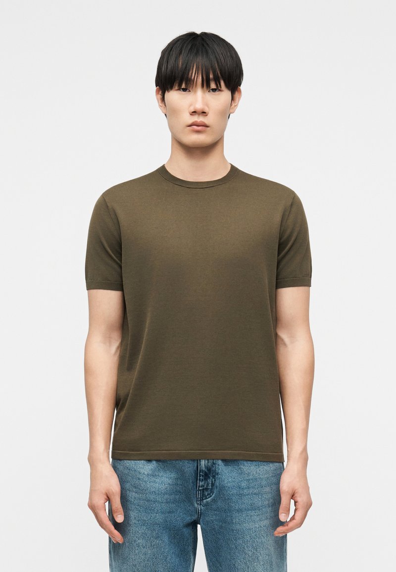 Νέος άνδρας με ίσια μαύρα μαλλιά που φοράει ένα χακί κοντομάνικο μπλουζάκι και μπλε τζιν, στέκεται μπροστά σε ένα απλό φωτεινό φόντο.