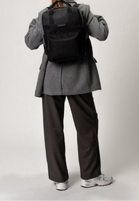 Osoba ubrana w szarą marynarkę, czarny plecak, ciemne spodnie o szerokich nogawkach oraz białe trampki, stojąca na tle jasnego, jednolitego tła.