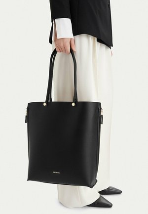 Persona con blazer negro y pantalones blancos sosteniendo un gran bolso tote de cuero negro con asas dobles y costados con cremallera.