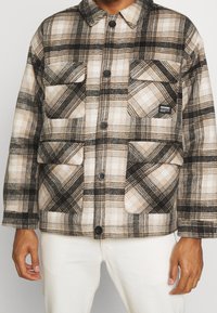 Rutig flanelljacka i beige, svart och creme. Har fyra framfickor, knäppning med knappar och en avslappnad passform. Mjuk textur.