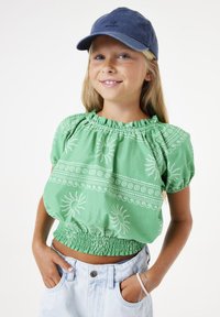Blusa de algodón verde con cintura elástica, con estampados de sol blancos y escote fruncido. Combinada con pantalones cortos de mezclilla azul claro.