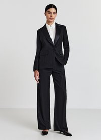 Costume noir à rayures avec revers en satin, veste cintrée et pantalons à jambes larges, porté sur une chemise à col blanc. Tissu lisse et structuré.