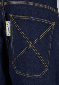 Jean en denim bleu foncé avec des coutures jaunes, présentant un motif en X distinctif sur la poche arrière et une étiquette "homeboy" attachée.