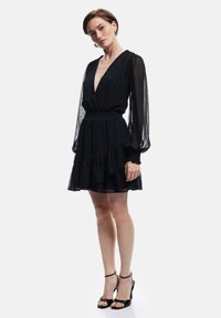 Schwarzes Kleid mit durchsichtigen, langen Ärmeln, tiefem V-Ausschnitt und gerafftem Saum. Verfügt über einen strukturierten Stoff mit dezentem Punktmuster. Kombiniert mit schwarzen Absätzen.