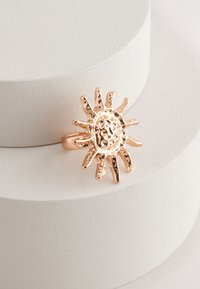 Gold-toned ring featuring a sun design with textured rays and a central circular pattern, set on a smooth light-colored surface.