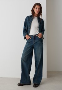 Dark denim jacket with buttons, paired with high-waisted wide-leg jeans. Both feature a casual fit and medium blue wash, set against a light background.