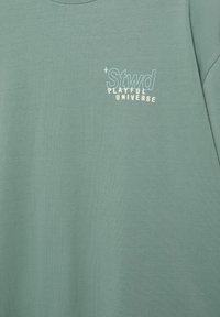 Material verde cu textul "Stwd" într-un font mare conturat și "PLAYFUL UNIVERSE" în litere mici drepte dedesubt, pe zona superioară a pieptului.