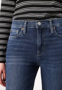 Blue denim jeans with a slim fit, featuring five pockets, a button closure, and subtle stitching details. Paired with a black and white striped top.