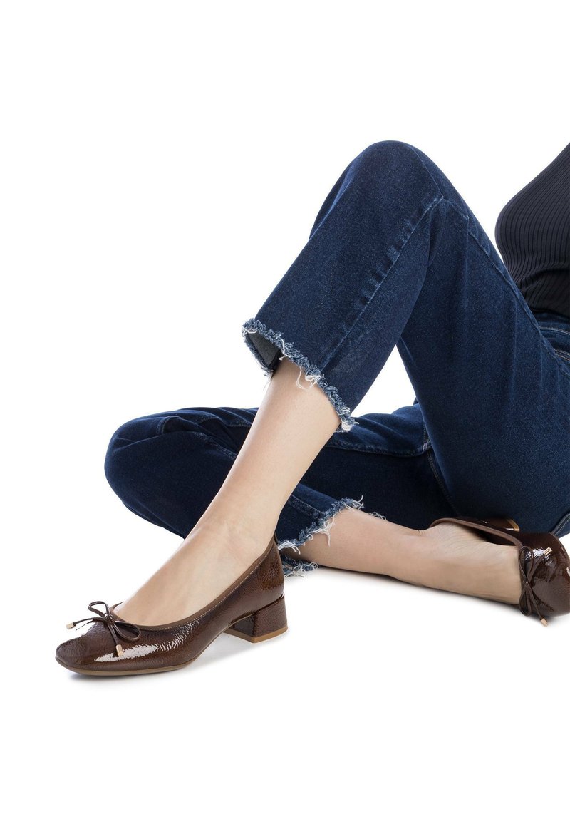 Bailarinas de cuero marrón con un pequeño tacón bajo, que presentan un detalle de lazo y un acabado texturizado. Llevadas con jeans azules deshilachados.