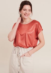 Glanzend roestkleurig T-shirt met korte mouwen en een relaxte pasvorm, gecombineerd met een crèmekleurige rok met knopen aan de voorkant en zakken.