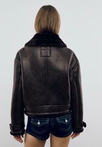 Giacca di pelle marrone con ampio colletto di pelliccia, dotata di schiena strutturata, maniche a polsino e una texture liscia. Indossata con pantaloni corti di jeans.