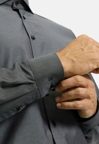 Graues Hemd mit Knöpfen und kleinem strukturiertem Muster, ausgestattet mit schwarzen Knöpfen und umgeschlagenen Ärmeln. Glatte Stoffqualität mit leichtem Glanz.