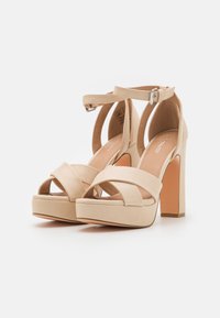 Beżowe sandały na wysokim platformowym obcasie z krzyżującymi się paskami, paskiem wokół kostki i kwadratowym obcasem. Gładki materiał o matowym wykończeniu.