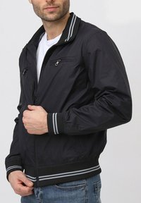 Veste bomber noire avec fermeture éclair à l'avant, col et poignets côtelés, et deux poches sur la poitrine avec fermeture éclair. Présente des accents rayés gris sur le col.