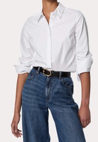 Hvid skjorte med krave og knapper, oprullede ærmer; parret med højtaljede blå denimjeans med et sort bælte med en guldbuckle.