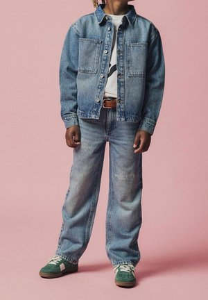 Lapsi, jolla on sininen farkkutakki ja farkut ruskealla vyöllä ja vihreät tennarit, seisoo vaaleanpunaisen taustan edessä.