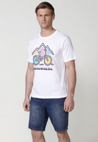 White cotton T-shirt featuring a colorful bicycle graphic and mountain design. Worn with blue denim shorts, presenting a casual look.