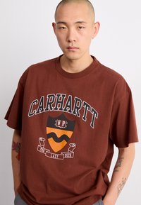 Brązowa bawełniana koszulka z grafiką w motywie tarczy, z napisem "CARHARTT" oraz "THE LAZY DUCK". Krótkie rękawy, luźny krój.
